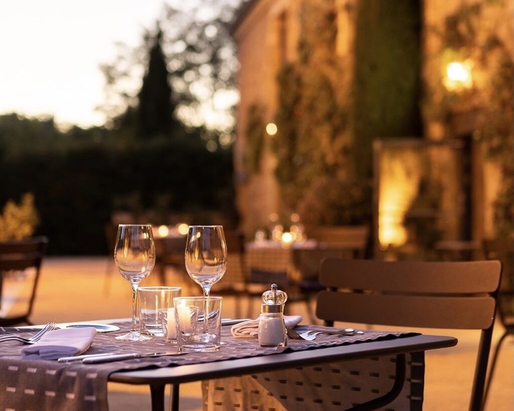 Une table à manger installée à l’extérieur, agrémentée de verres à vin, d’un salier et d’une bougie, avec un mur de pierre et des arbres en arrière-plan.