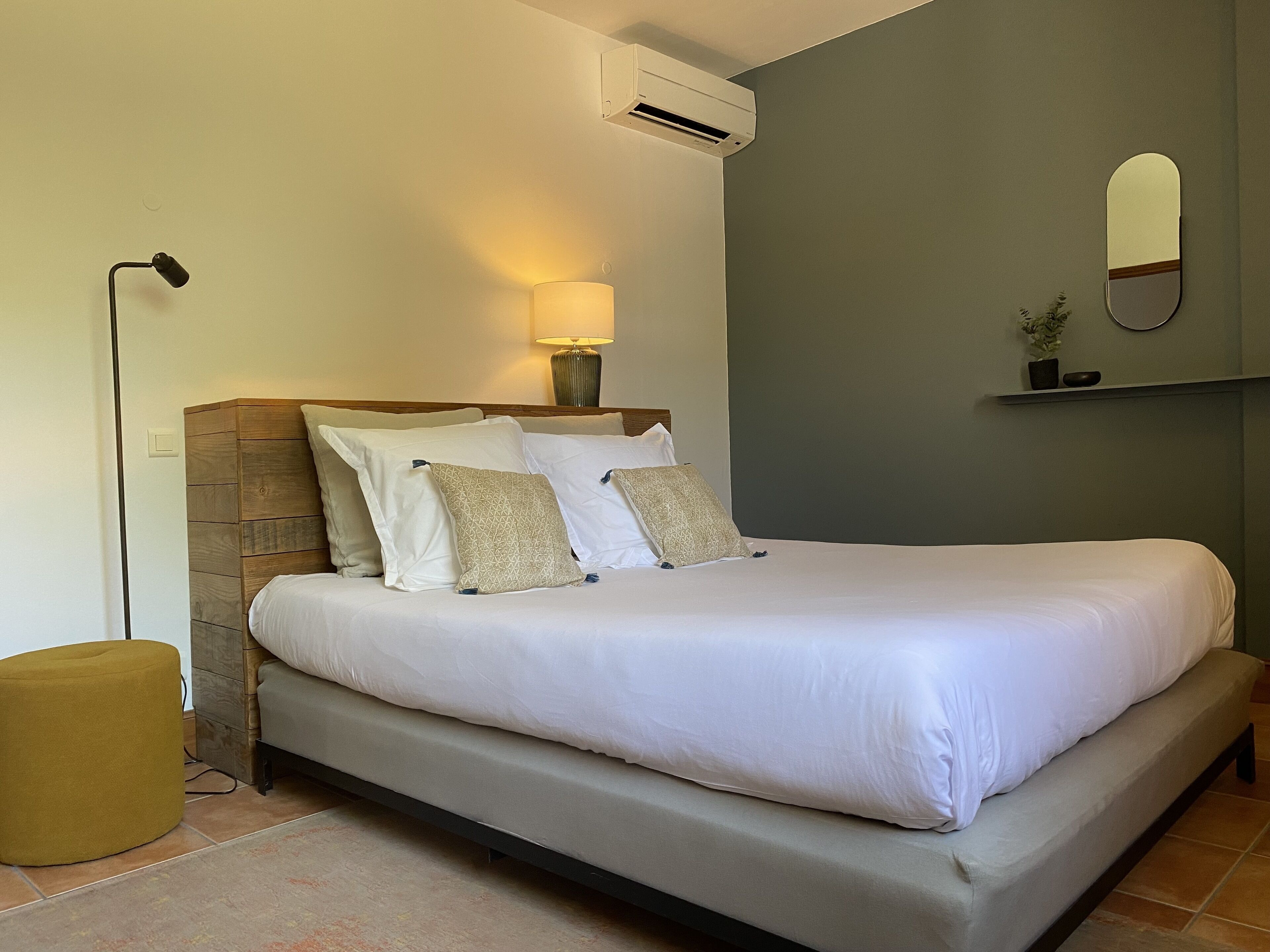 A neatly made bed with white linens and several pillows, a bedside lamp, and an air conditioning unit on the wall.