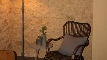 A room with a stone wall, a wicker chair with a cushion, a small table with a lamp, and a framed picture on the wall.