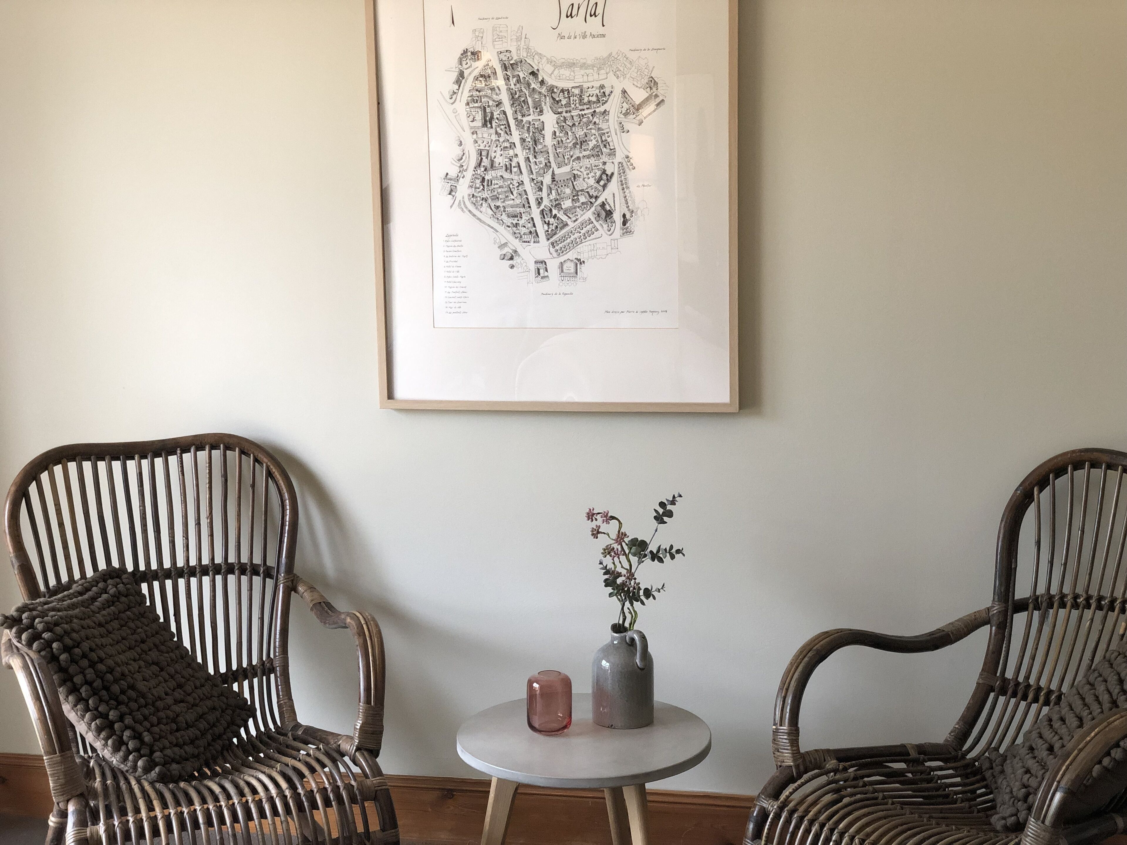 A framed map on the wall, two wicker chairs, a small round table with a vase and a candle.