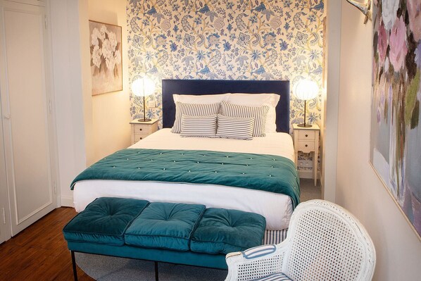 Une chambre à coucher avec un lit, des lampes de chevet, un banc recouvert d’un tissu vert et une chaise blanche.