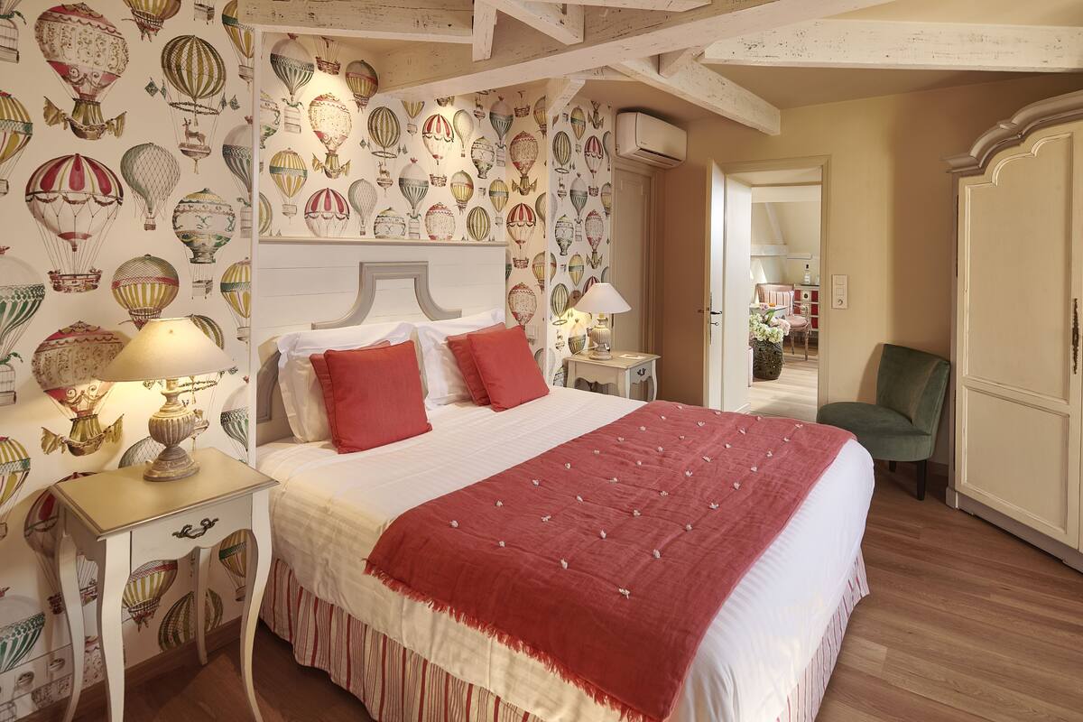 Ein Schlafzimmer mit einem großen Bett, zwei Nachttischen mit Lampen, einem grünen Sessel und einer Wand mit einem Heißluftballon-Muster.