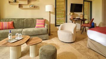 Habitación de hotel con sofá, sillón, reposapiés, una mesita con bandeja, lámpara, televisión y cama.