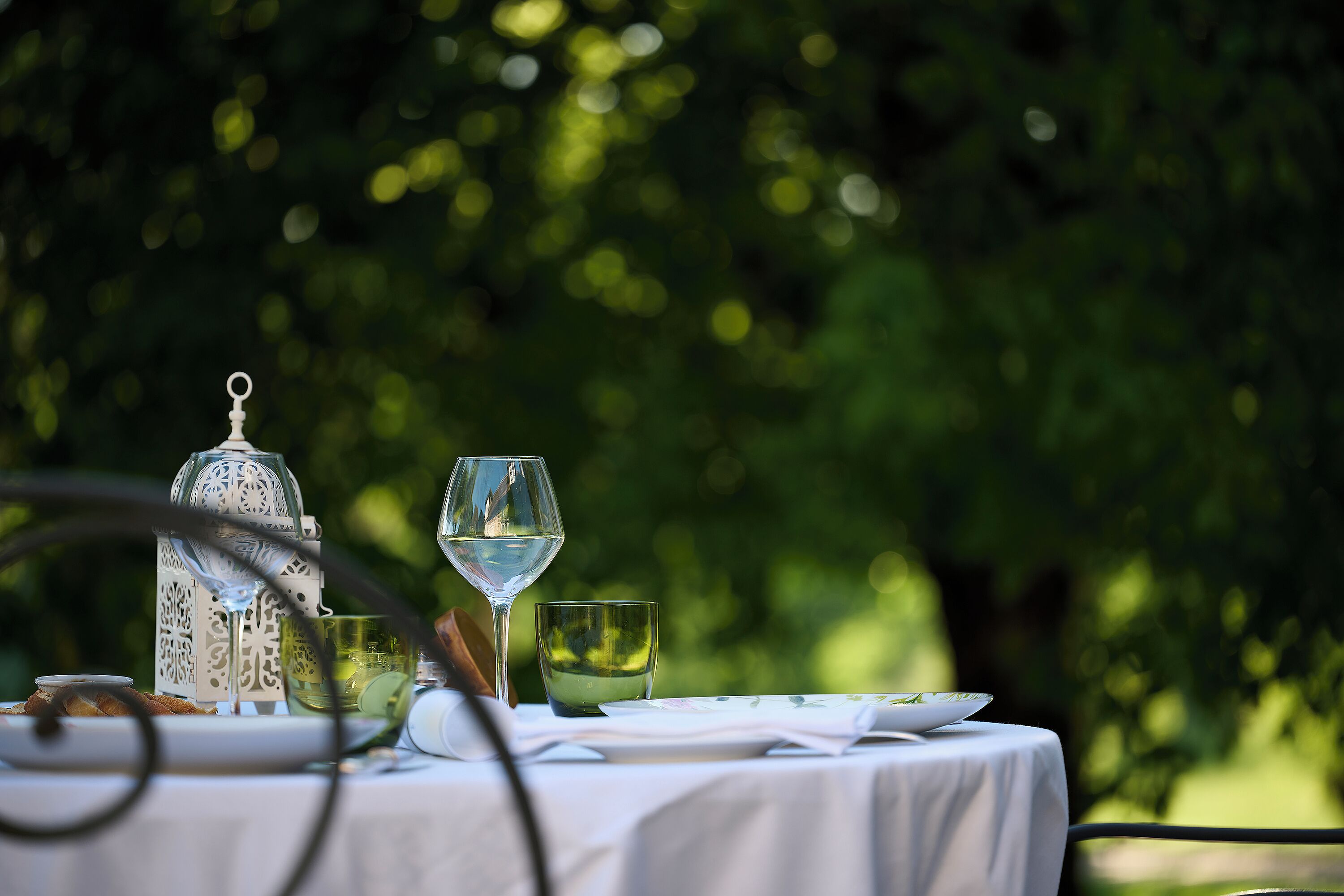 Une table dressée à l’extérieur, recouverte d’une nappe blanche, avec un verre de vin blanc, un verre d’eau, une lanterne décorative et une assiette de nourriture.