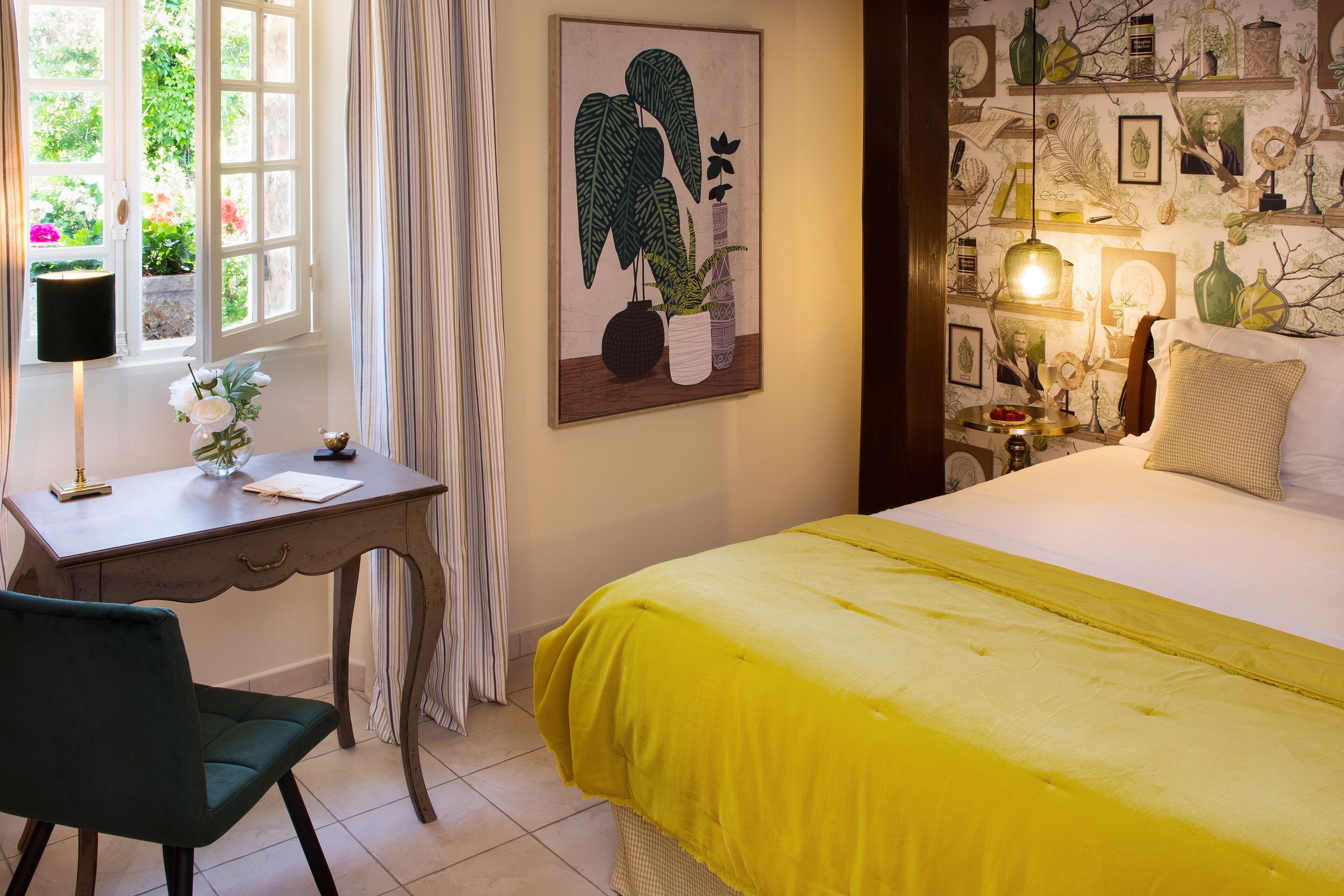 Un dormitorio con cama, escritorio y silla. Se observa una ventana con cortinas, una pared adornada con cuadros enmarcados, y una mesita de noche con lámpara.