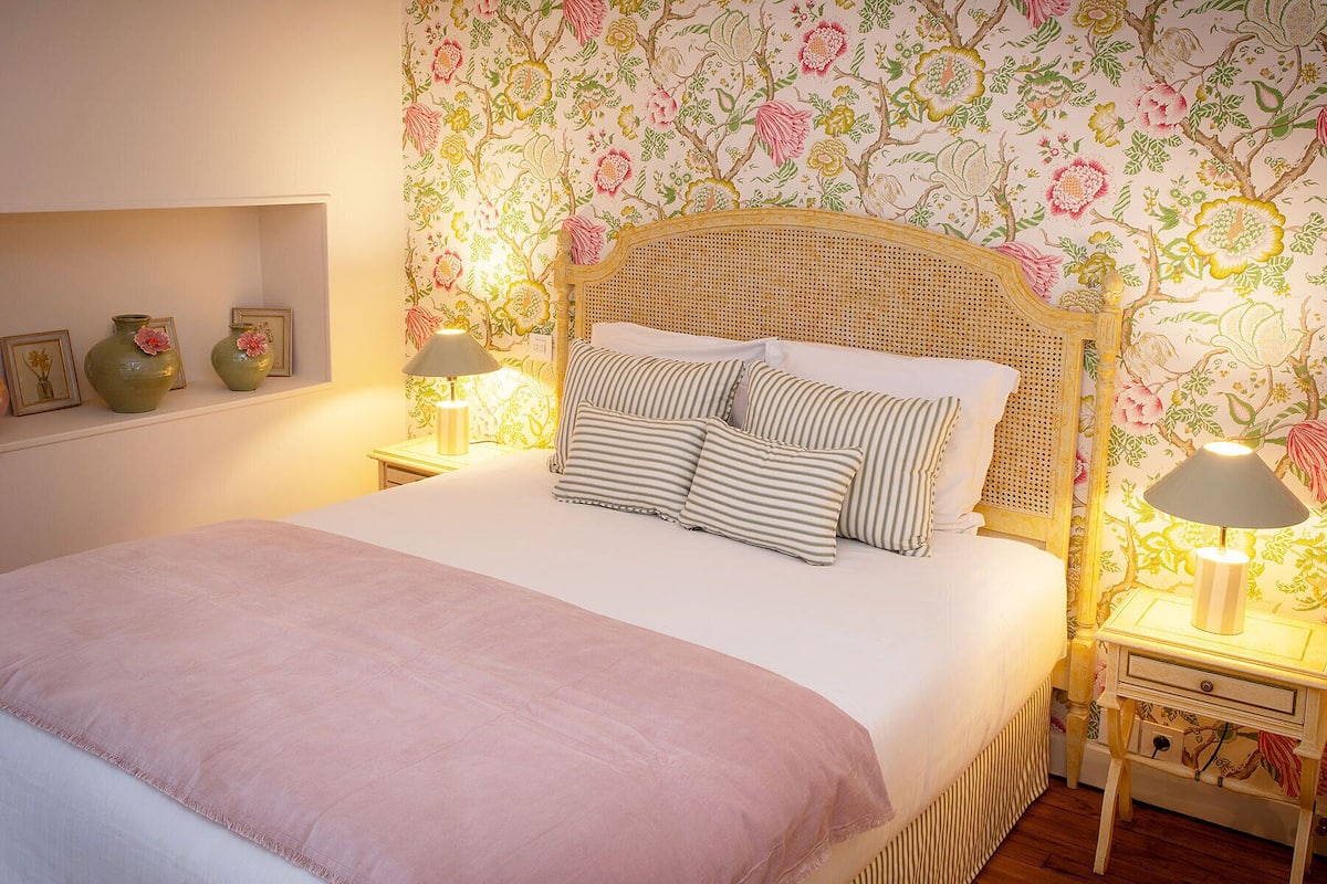 Ein ordentlich bezogenes Bett mit Kissen in Streifenoptik, eine Nachttischlampe und eine Wand mit einem floralen Muster.