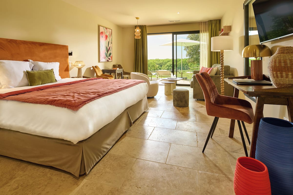 Ein Hotelzimmer mit einem großen Bett, einem Schreibtisch mit Lampe, einem Sessel und Blick ins Freie durch ein großes Fenster.