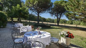 An outdoor dining area with round tables and chairs, surrounded by trees and a clear sky.