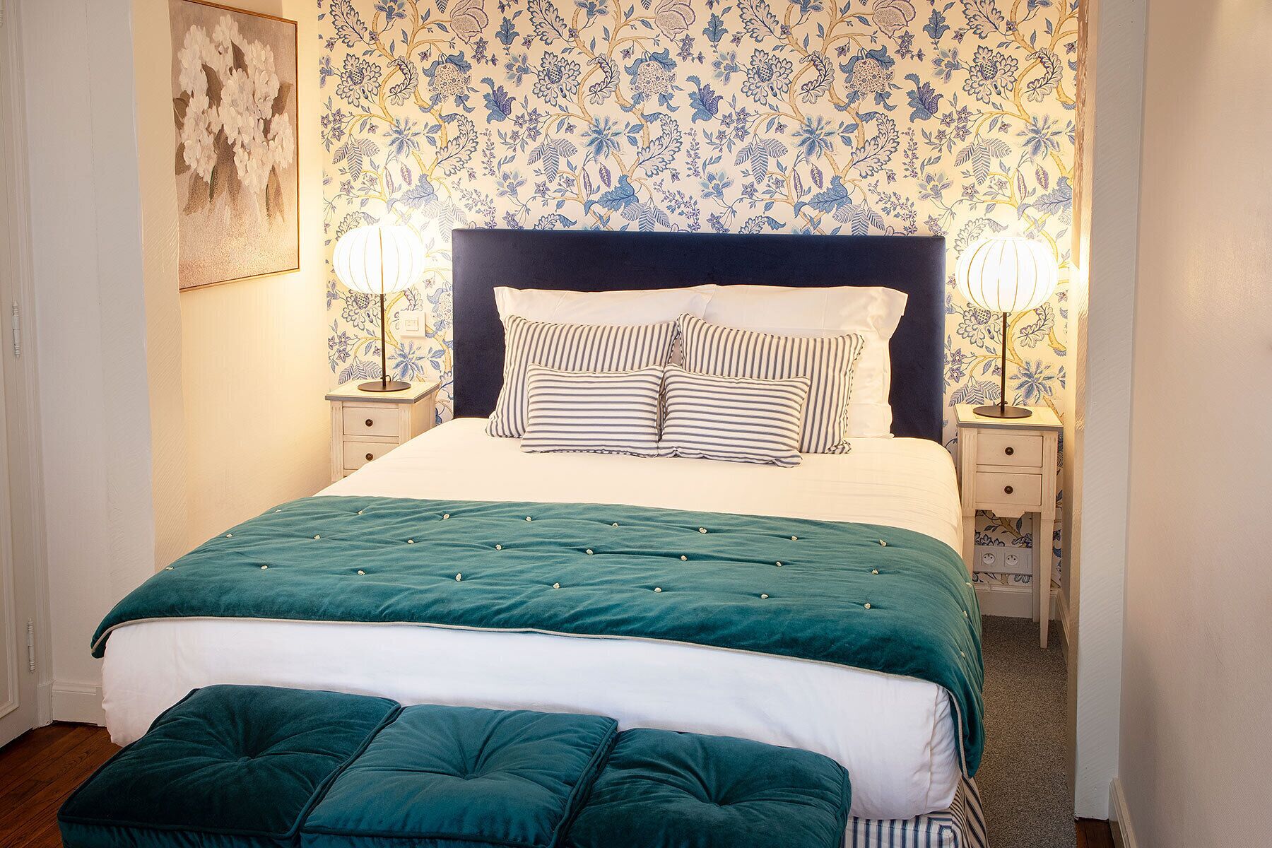 A neatly made bed with a striped pillow set, a teal bedspread, and a matching ottoman. There are two bedside lamps on nightstands with floral wallpaper.