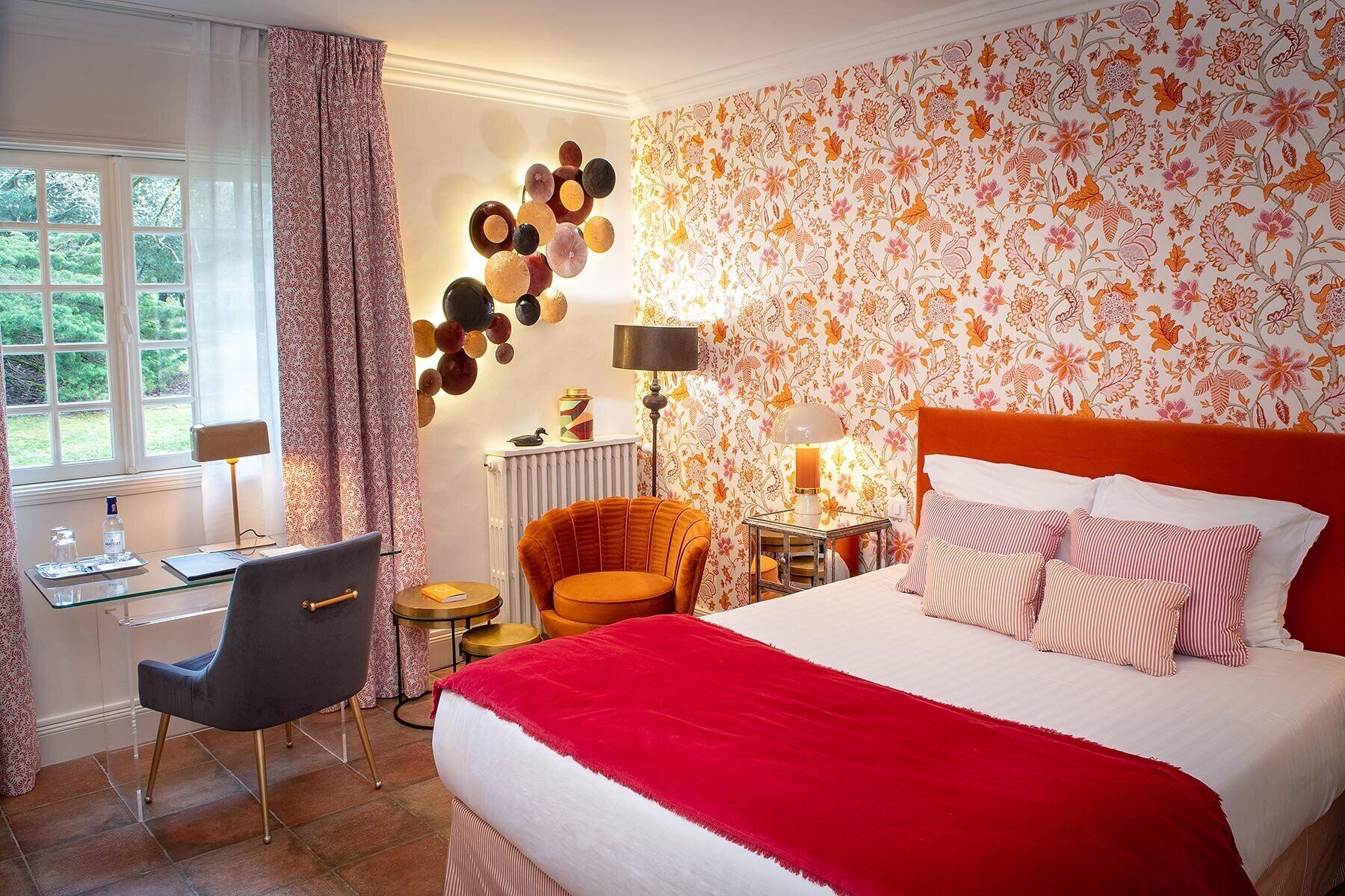 Een hotelkamer met een bed, een bureau, een stoel, een lamp en een bloemenpatroon op de muur.