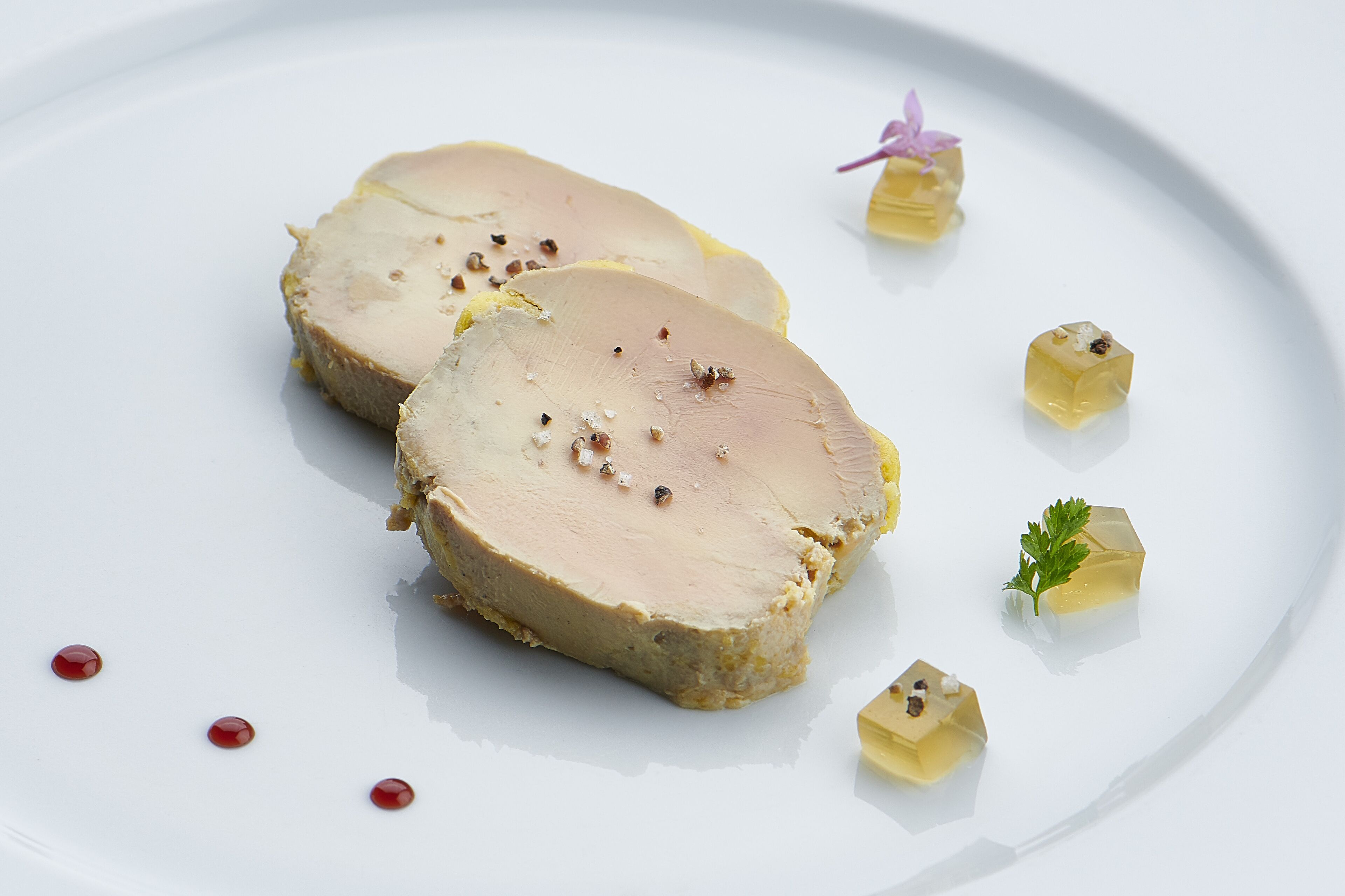 A white plate with slices of meat and small cubes of a yellowish substance, possibly a dessert, garnished with a purple flower and parsley.