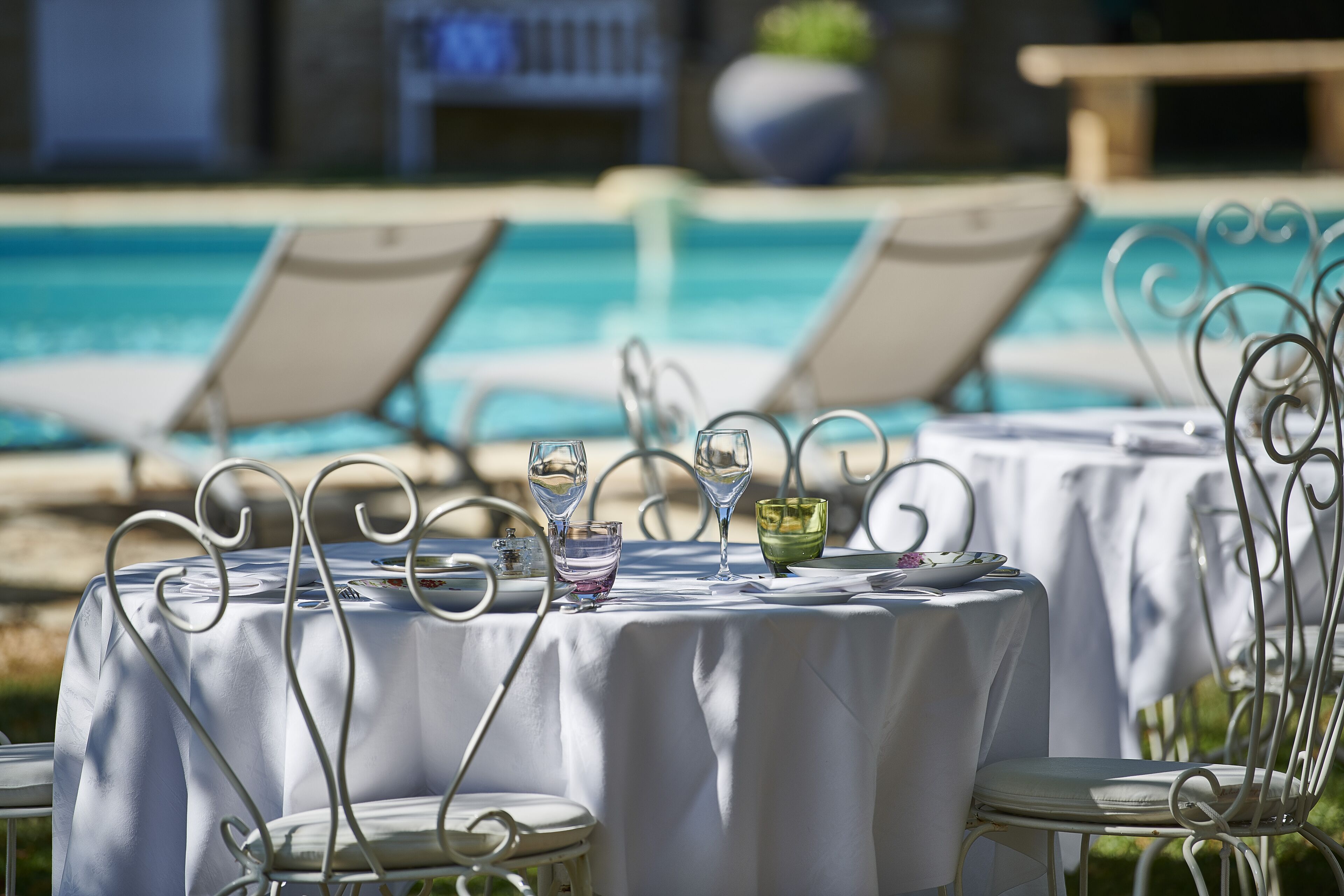 A poolside dining area with tables set for two, each with a metal chair and a white tablecloth.