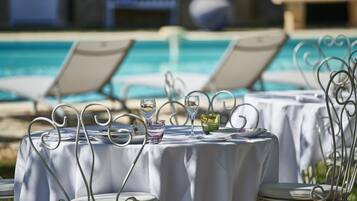 A poolside dining area with tables set for two, each with a metal chair and a white tablecloth.