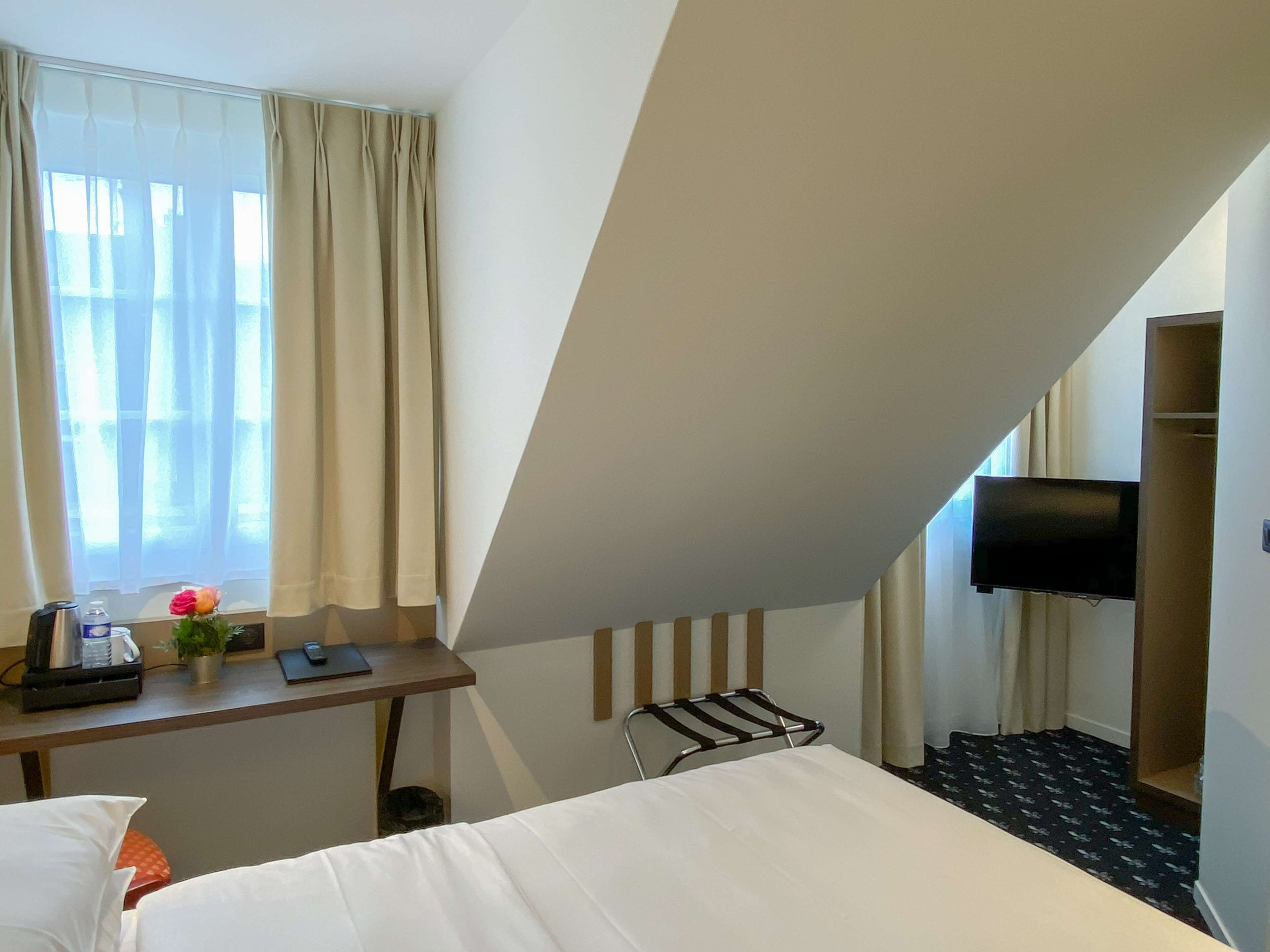A hotel room with a bed, a desk with a chair, a TV mounted on the wall, and a window with curtains.