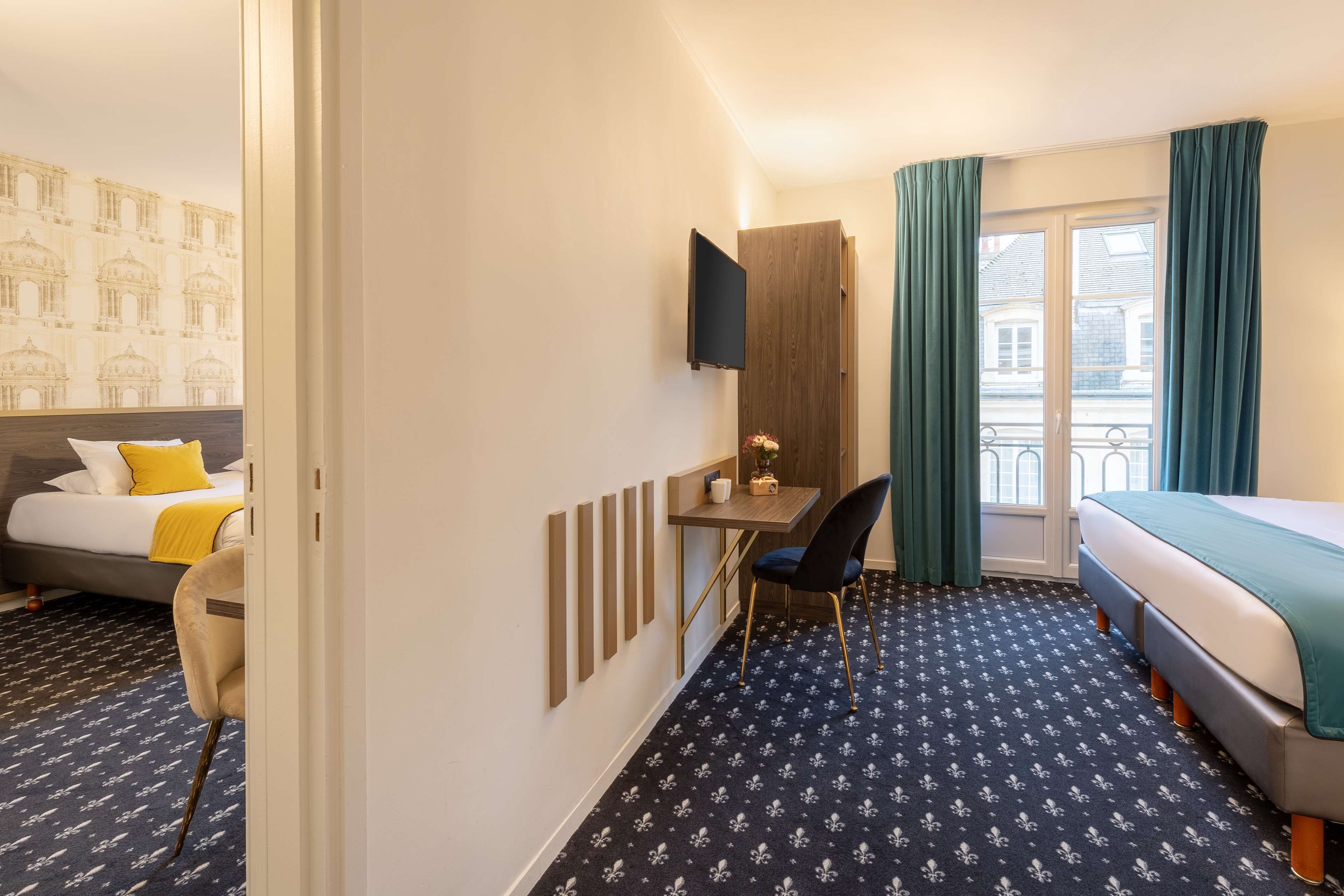 A hotel room with a bed, a desk with a chair, a TV mounted on the wall, and a window with curtains.