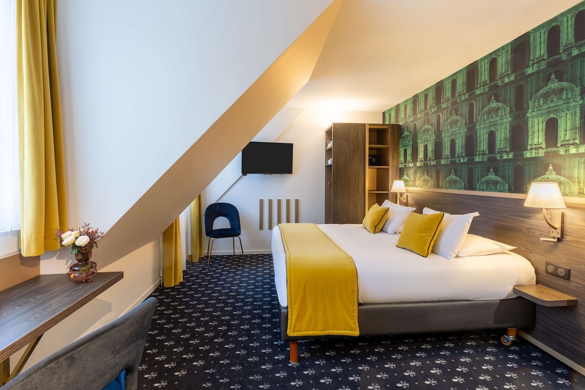 A hotel room with a bed, a desk, a chair, a TV, and a wall with a green architectural mural.
