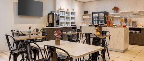 Un comedor moderno con mesas de mármol y sillas negras, una cafetera y un bufé con diversas opciones de comida.