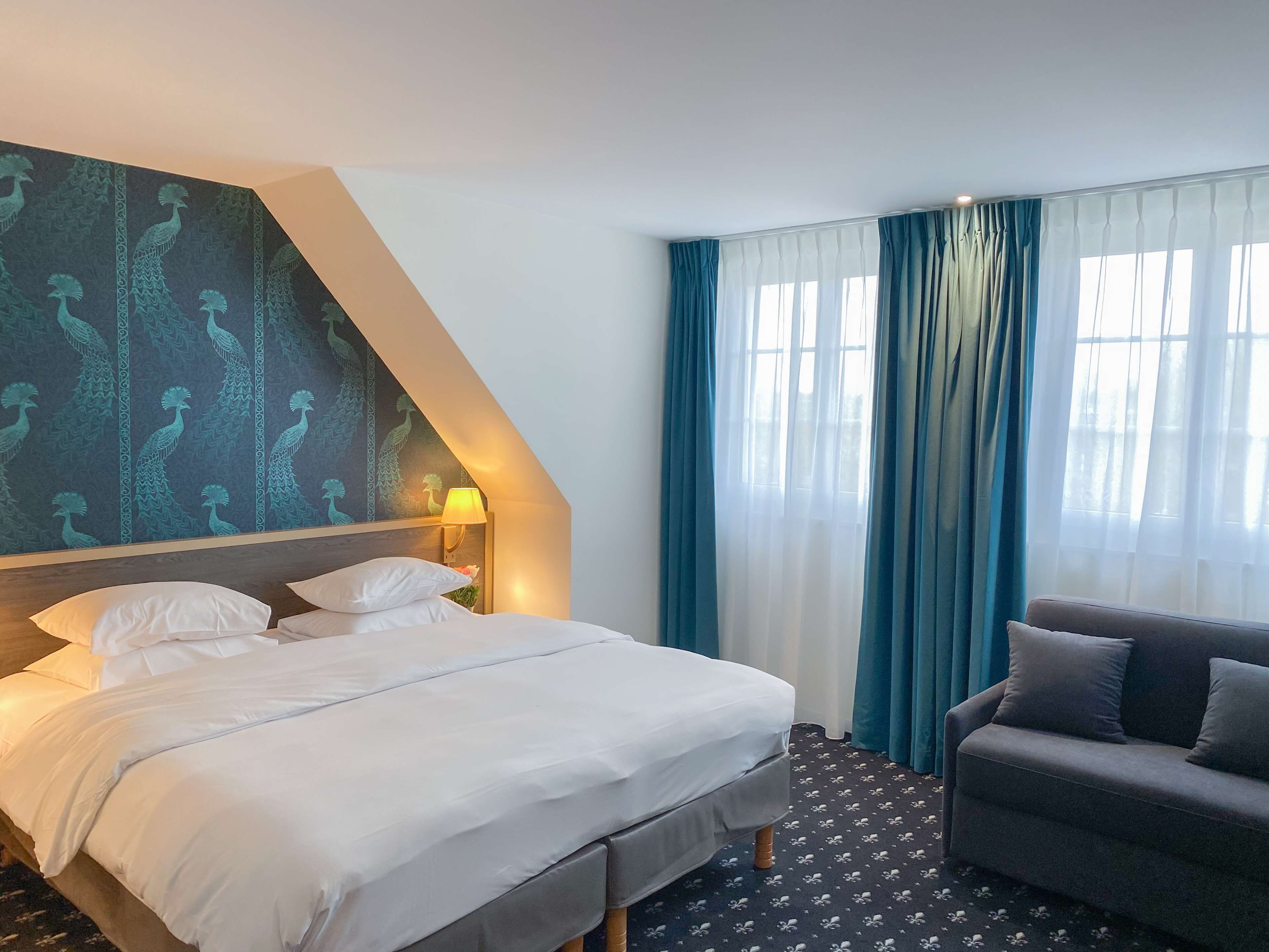A hotel room with a bed, a sofa, a lamp, and a window with curtains.