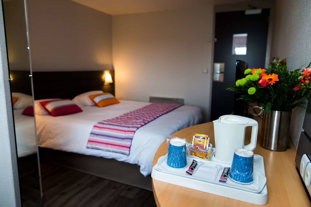 A hotel room with a bed, a bedside table with a tray, a coffee maker, a vase with flowers, and a nightstand.