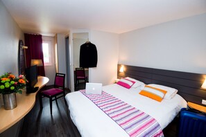 A hotel room with a bed, a desk with a laptop, a chair, a TV, and a vase of flowers.