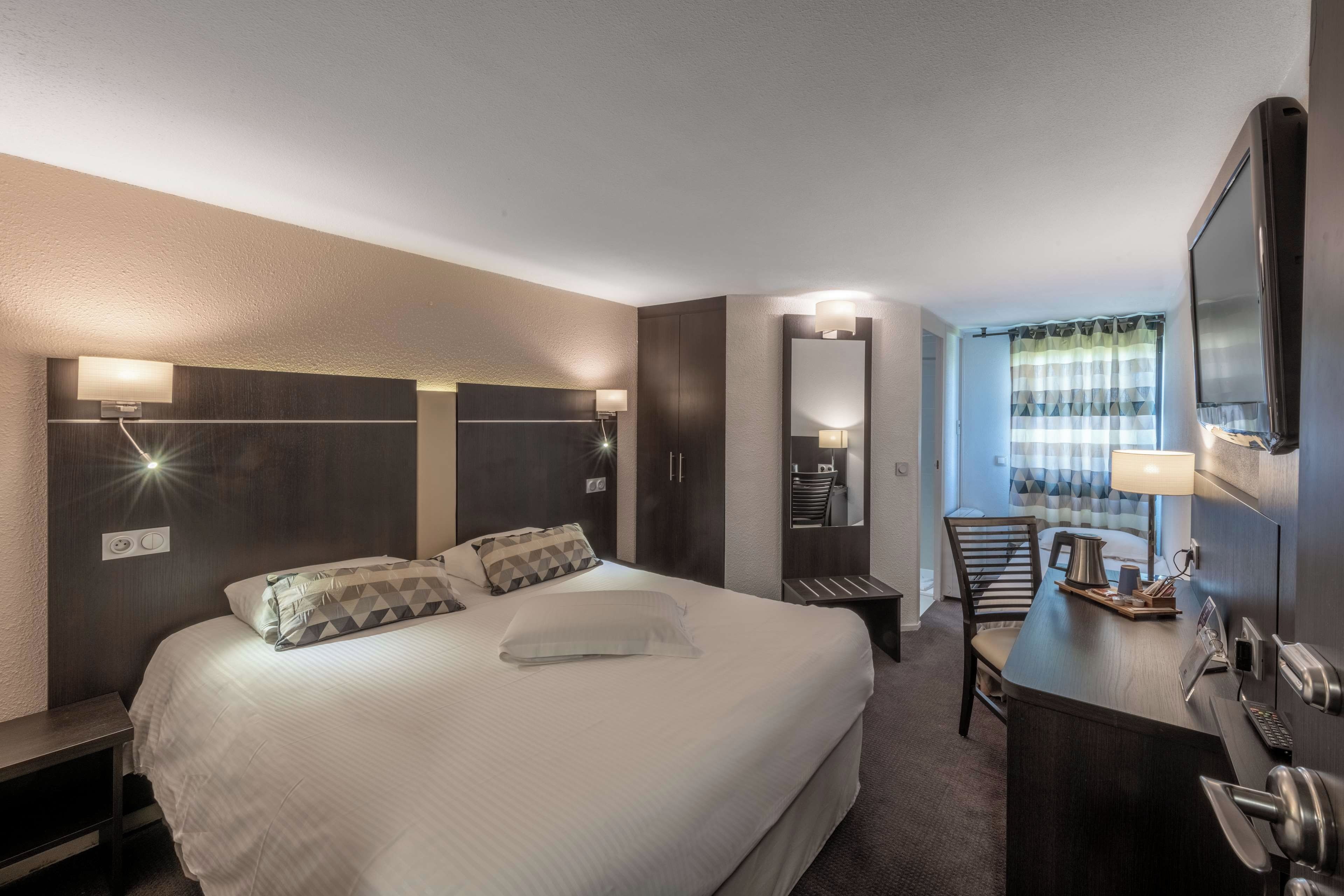 A hotel room with a large bed, a desk with a chair, a mirror, and a window with curtains.