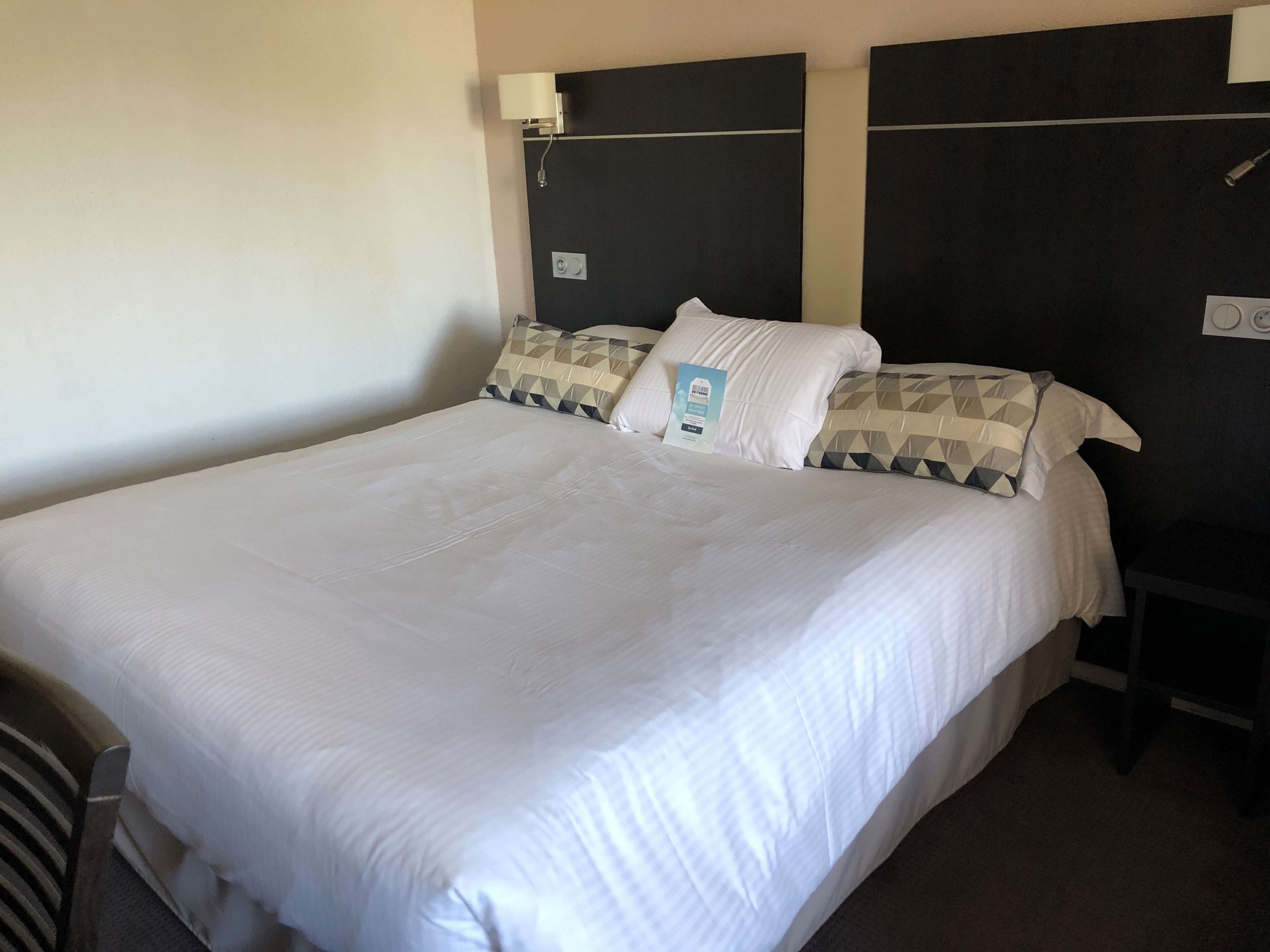 A neatly made bed with white bedding, a pillow with a geometric pattern, and a bedside table with a lamp.