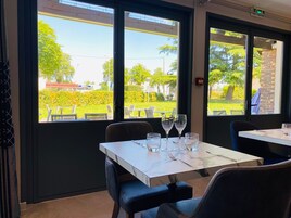 Comedor con mesa lista para dos personas, ventanas amplias con vista a un jardín y mobiliario exterior.