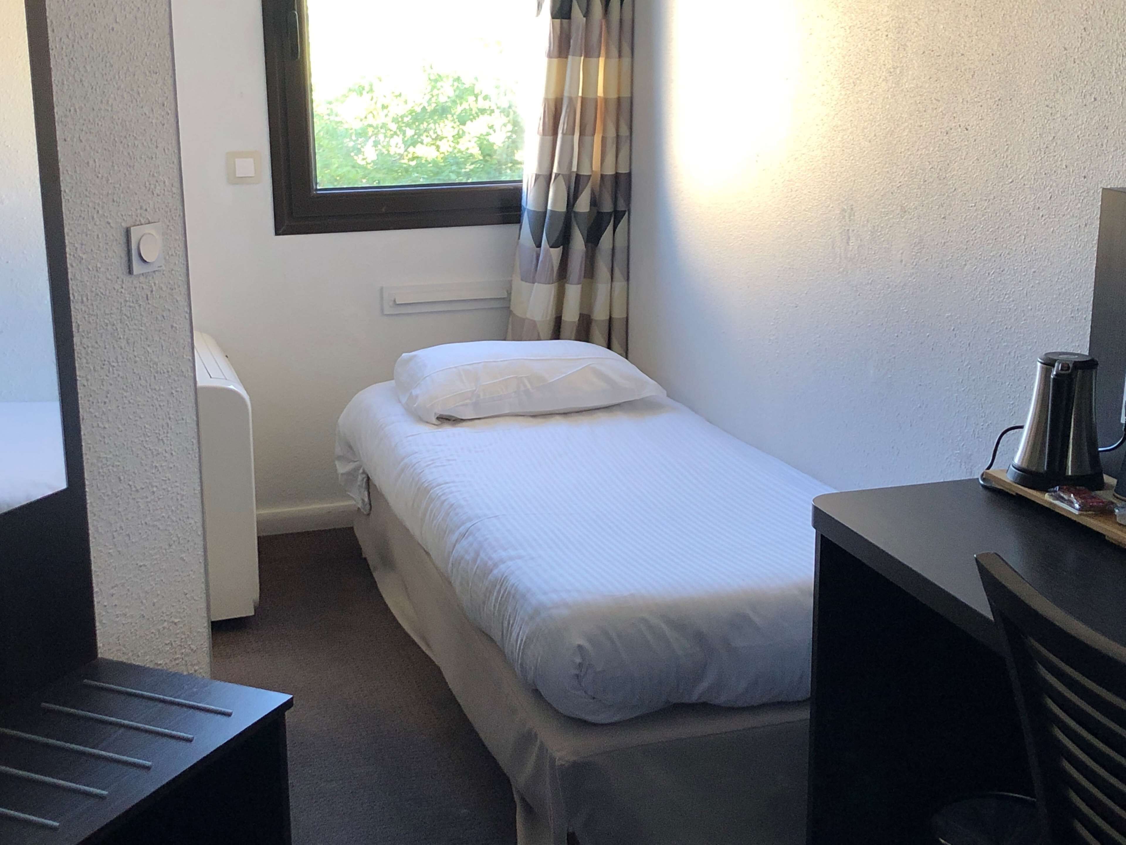 Ein kleines Zimmer mit einem Einzelbett, einem Fenster, einem Heizkörper, einem Schreibtisch und einem Wasserkocher.