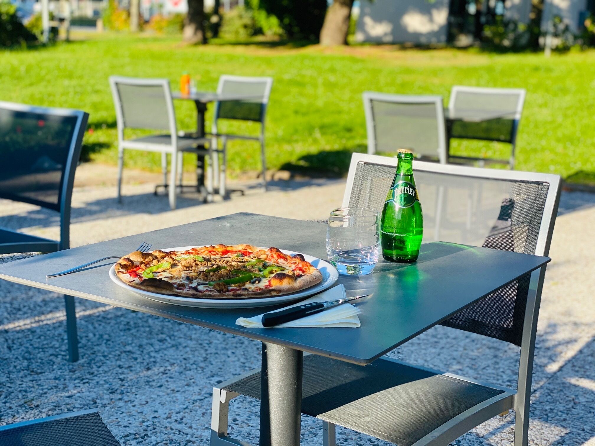 Una mesa de comedor al aire libre con pizza, una botella de agua Perrier y un vaso.