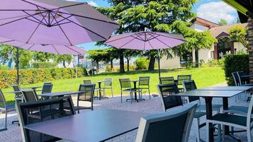 An outdoor seating area with tables and chairs, shaded by large umbrellas, and a view of a well-maintained garden and buildings in the background.