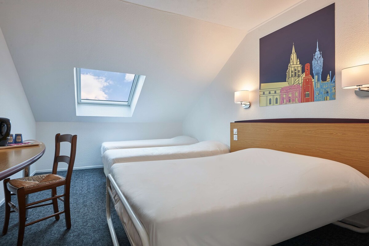 A hotel room with a single bed, a desk with a chair, a small window, and a cityscape painting on the wall.
