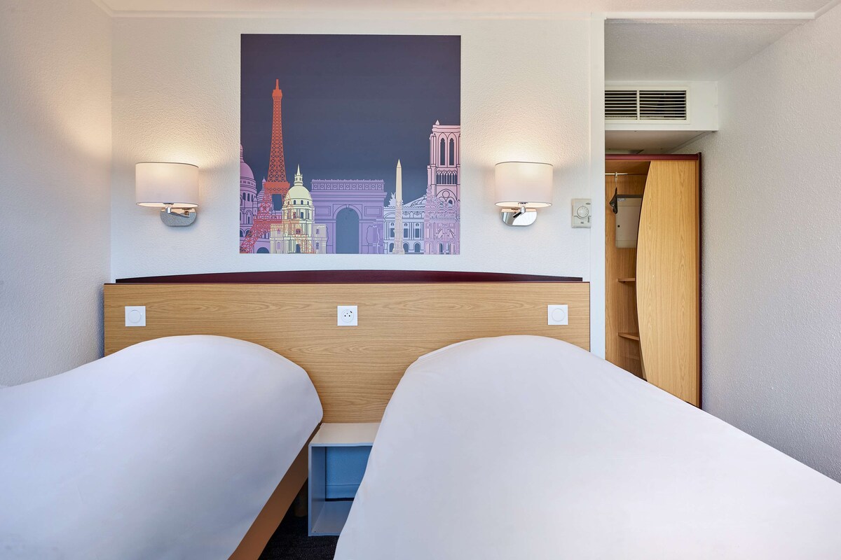 A hotel room with two single beds, a wall-mounted artwork of Parisian landmarks, and two wall-mounted reading lights.
