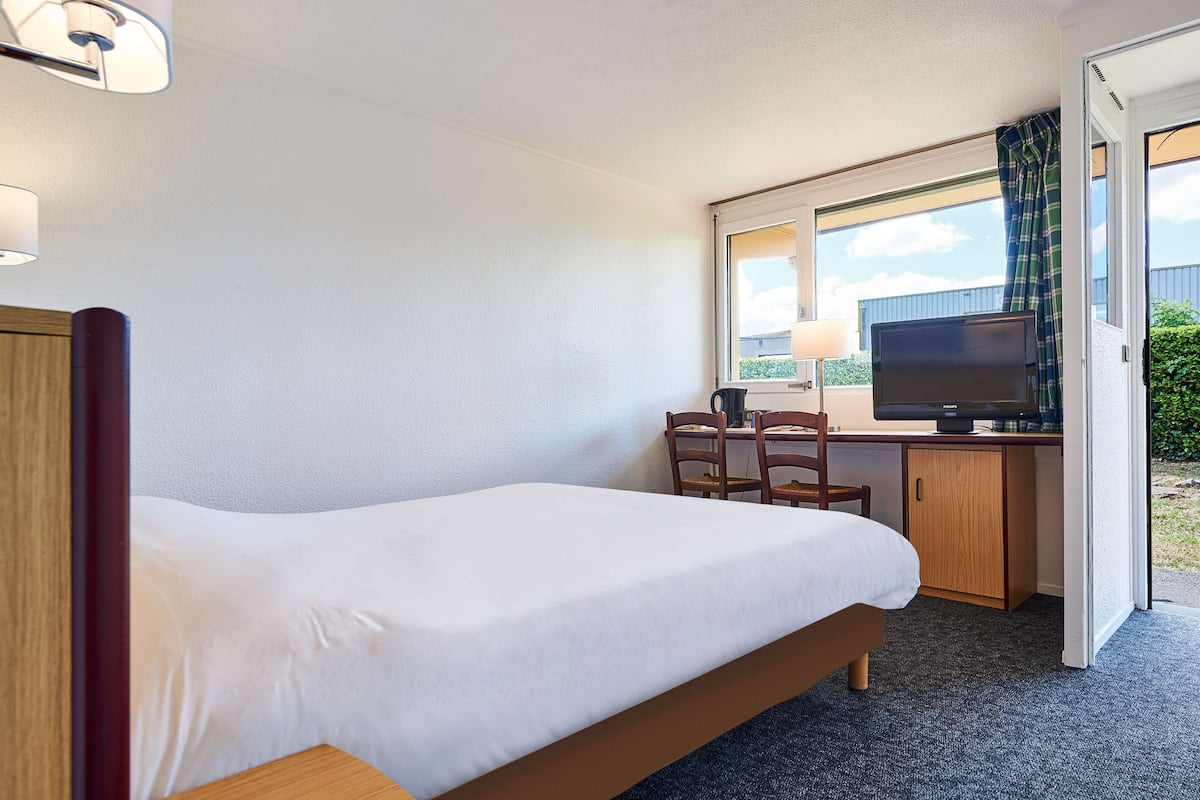 Une chambre d’hôtel avec un lit simple, un bureau équipé d’un ordinateur et d’une télévision, et une fenêtre avec des rideaux.