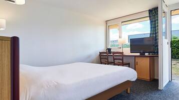 A hotel room with a single bed, a desk with a chair, a television, and a window with curtains.