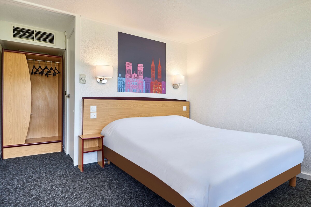 A hotel room with a bed, a wooden wardrobe, a coat rack, and a cityscape painting on the wall.