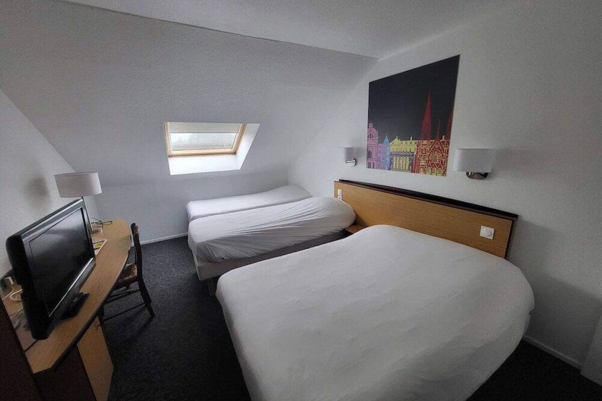 Une chambre d’hôtel avec un lit simple, un bureau avec un ordinateur, une télévision, une lucarne et un tableau accroché au mur.
