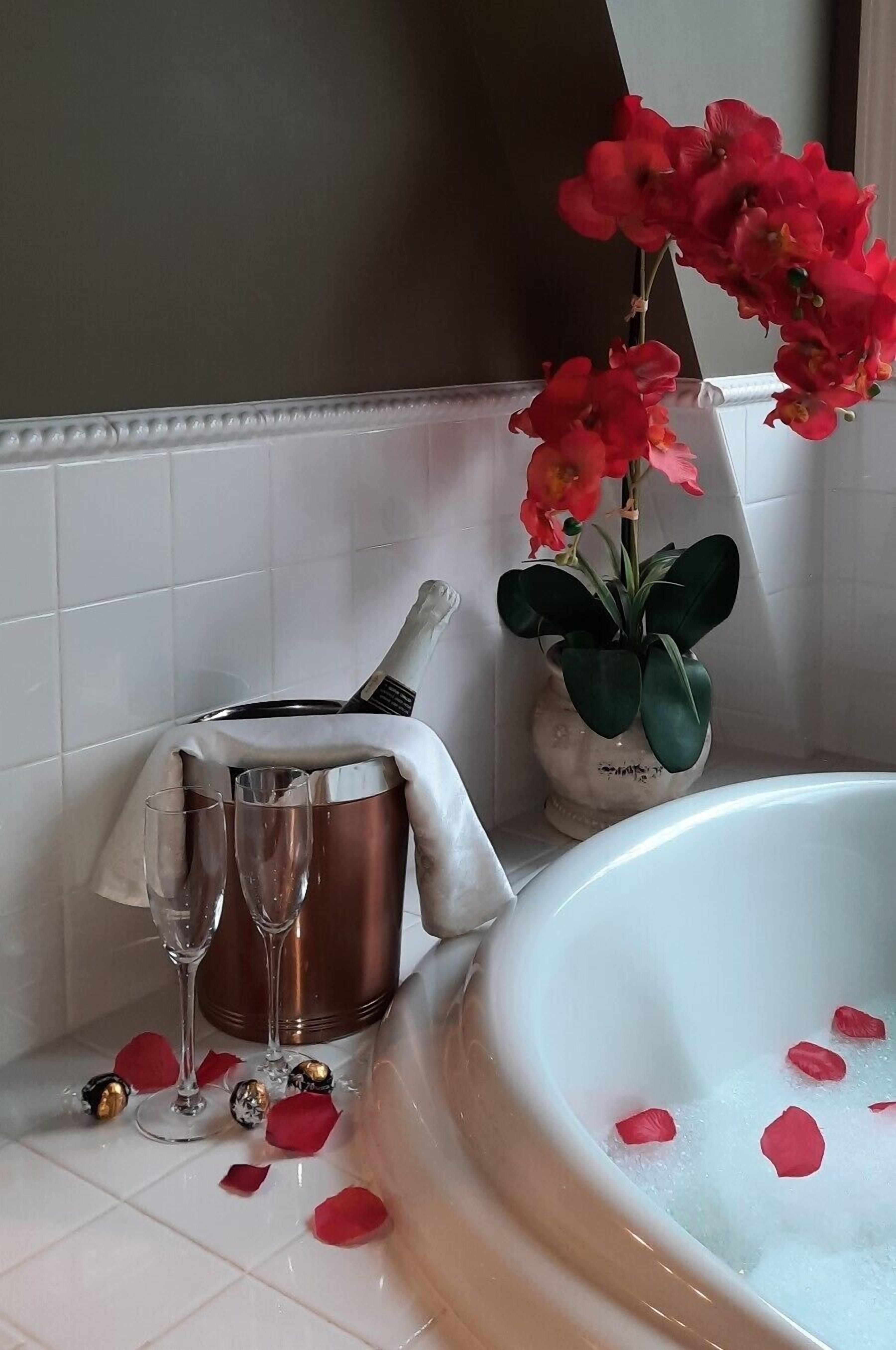 A bathroom with a bathtub filled with water and rose petals, a champagne bottle, two champagne flutes, and a potted plant with red flowers.
