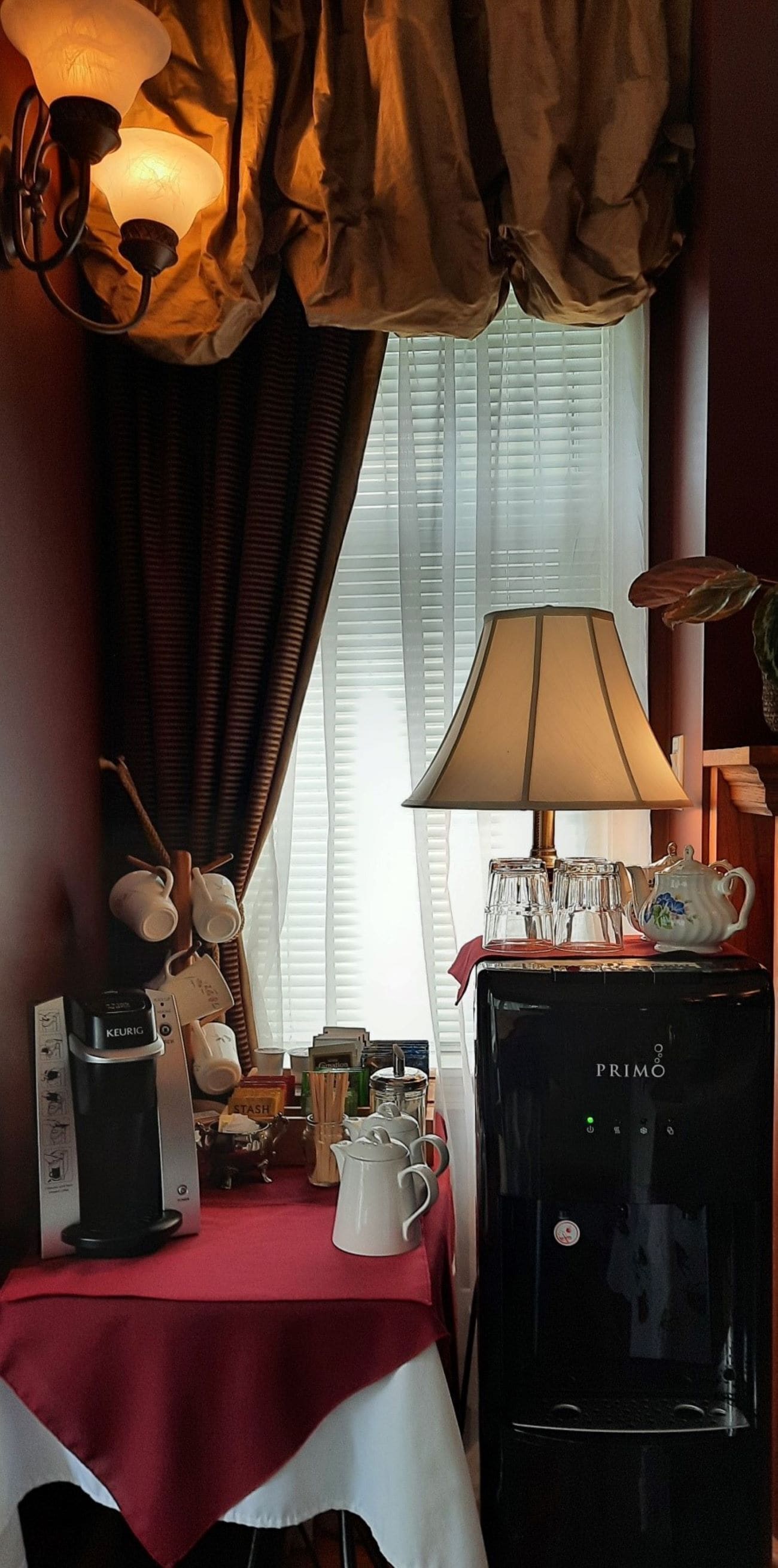 A hotel room with a coffee maker, a table with a red tablecloth, a lamp, and a window with curtains.