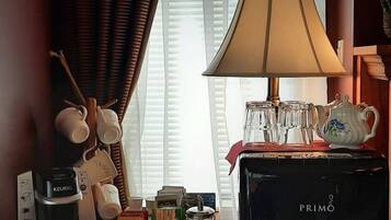 A hotel room with a coffee maker, a table with a red tablecloth, a lamp, and a window with curtains.