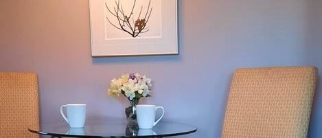 Een eetgedeelte met twee stoelen, een tafeltje, een vaas met bloemen en een lijst met een afbeelding aan de muur.