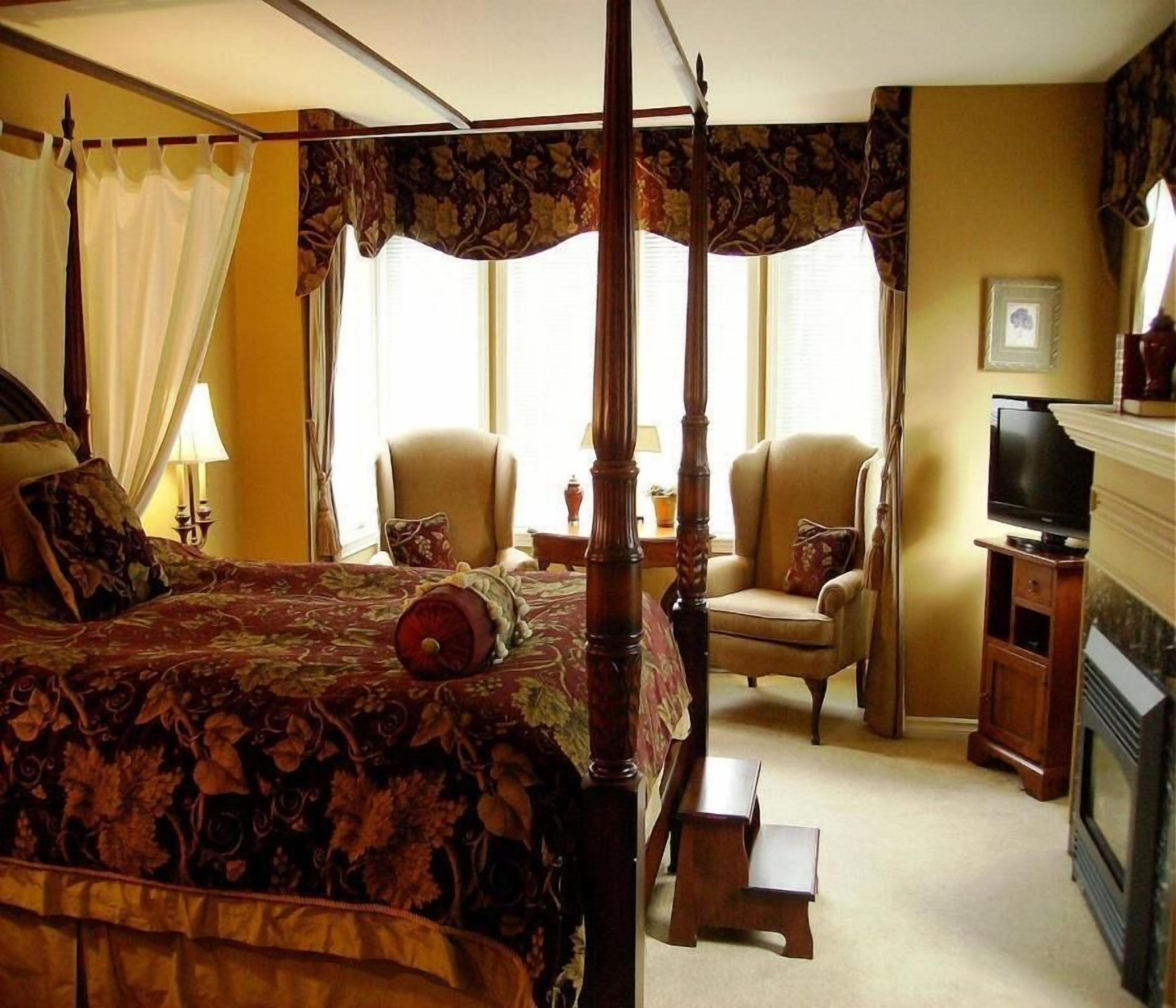 Ein Himmelbett mit floralem Überwurf, zwei Sessel, ein kleiner Tisch, ein Fernseher und ein Kamin.