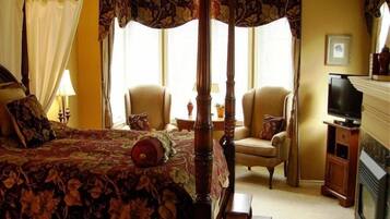 A four-poster bed with a floral bedspread, two armchairs, a small table, a television, and a fireplace.