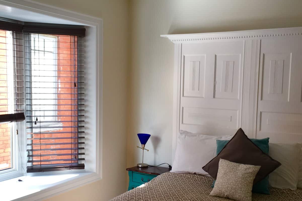 A bedroom with a bed, a nightstand, a lamp, a window with blinds, and a ceiling fan.