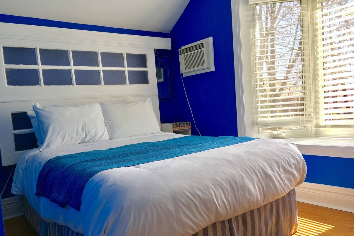 A bedroom with a bed, white headboard, blue walls, and a window with blinds.