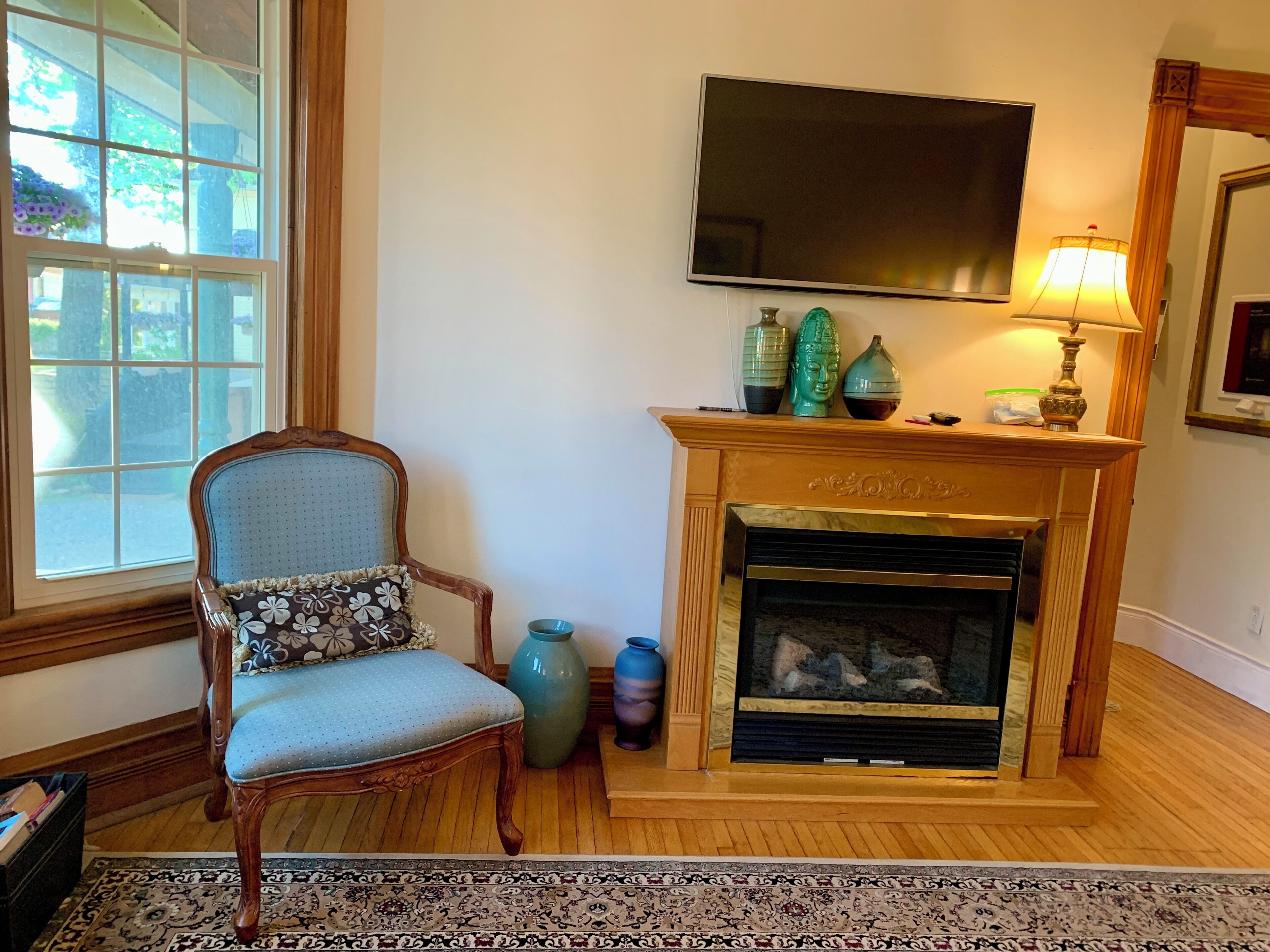 Une pièce avec une cheminée, un téléviseur fixé au-dessus, un fauteuil avec un coussin à motifs et une grande fenêtre.