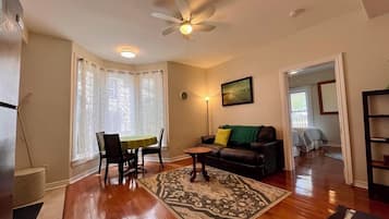 A living room with a dining table, a sofa, a patterned rug, a ceiling fan, and a painting on the wall.