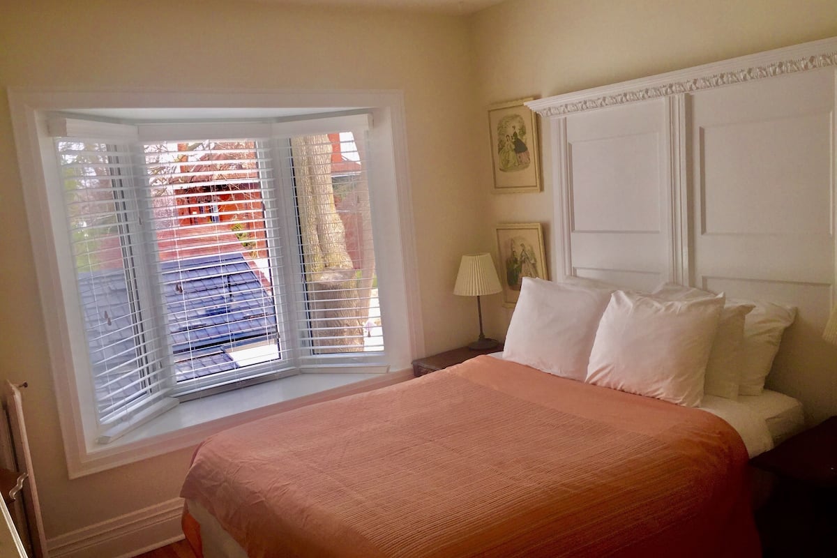 A bedroom with a large bed, a window with blinds, a nightstand with a lamp, and framed pictures on the wall.