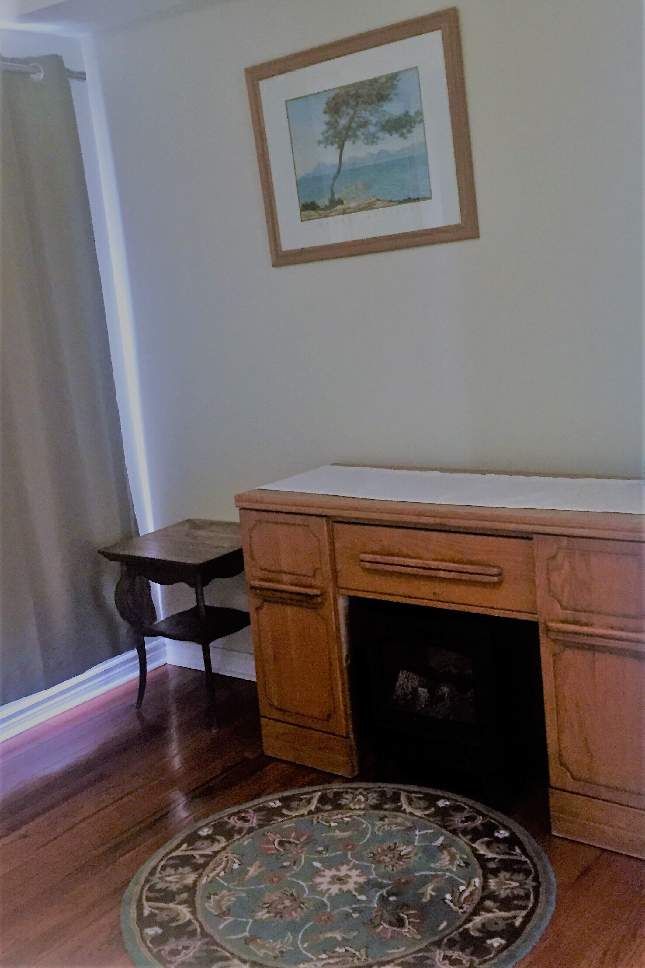 A room with a wooden desk, a small table, a patterned rug, and a framed picture on the wall.