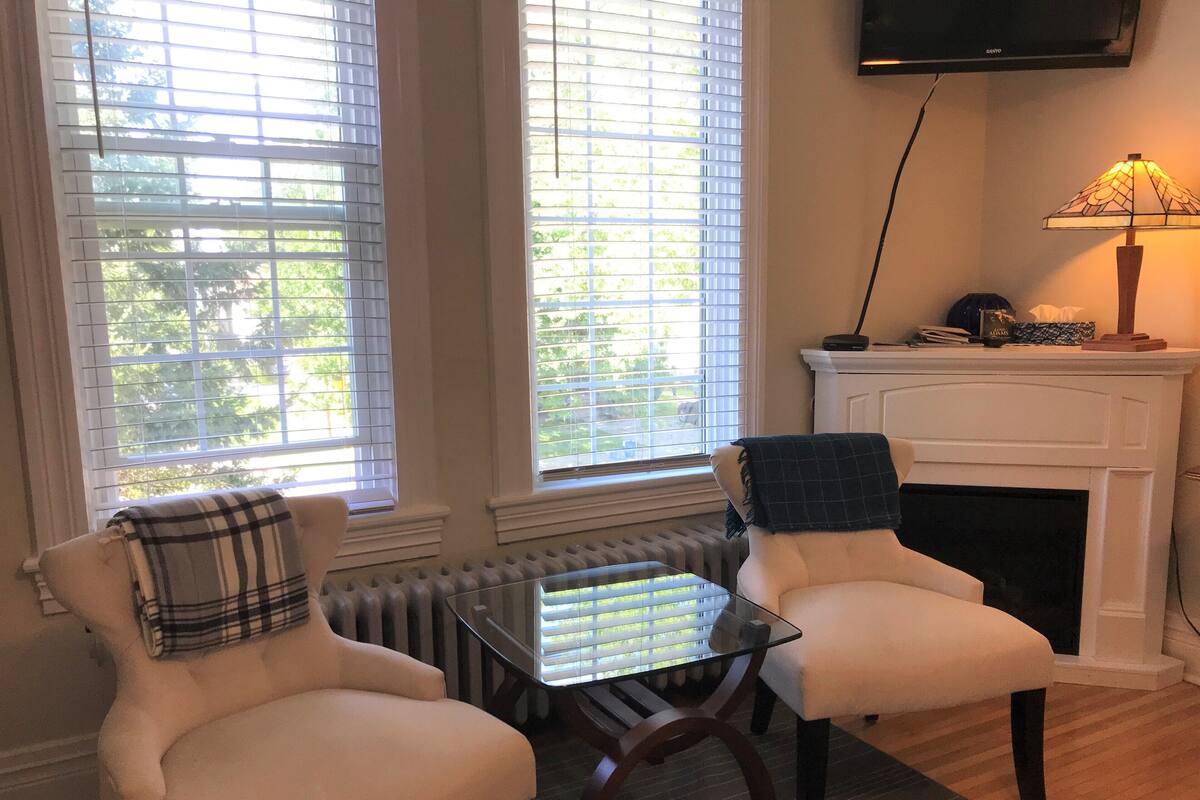 A cozy living room with two armchairs, a glass-top coffee table, a fireplace, and a television mounted on the wall.