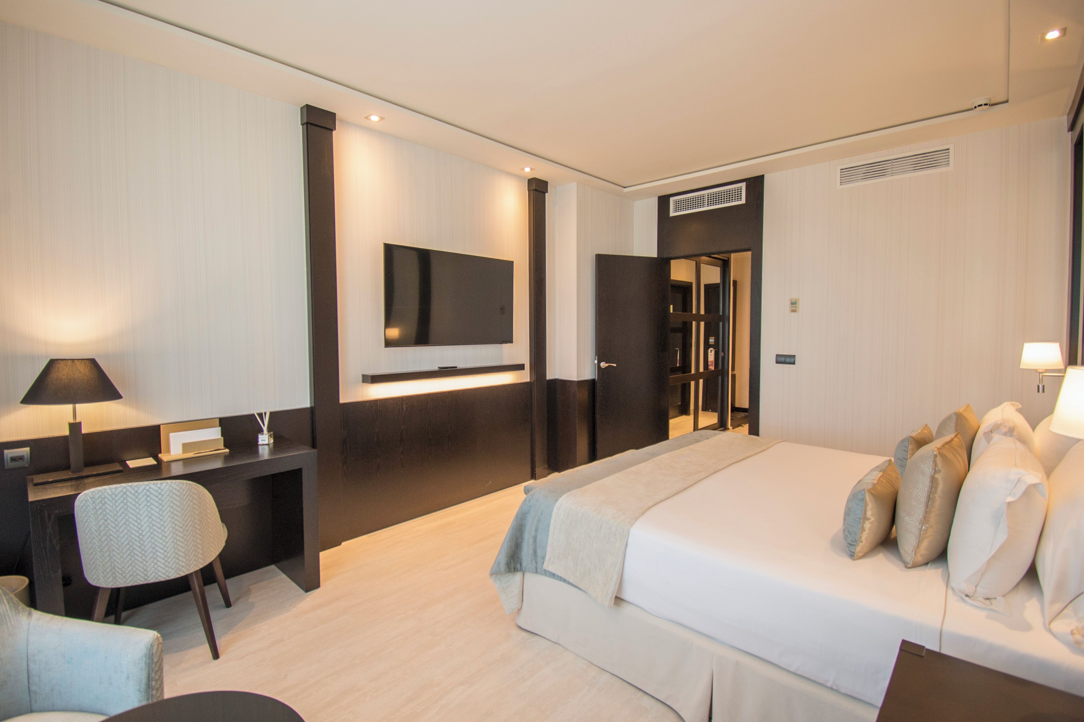 Una habitación de hotel moderna con una cama grande, un escritorio, una silla, un televisor montado en la pared y una puerta que da a otra habitación.